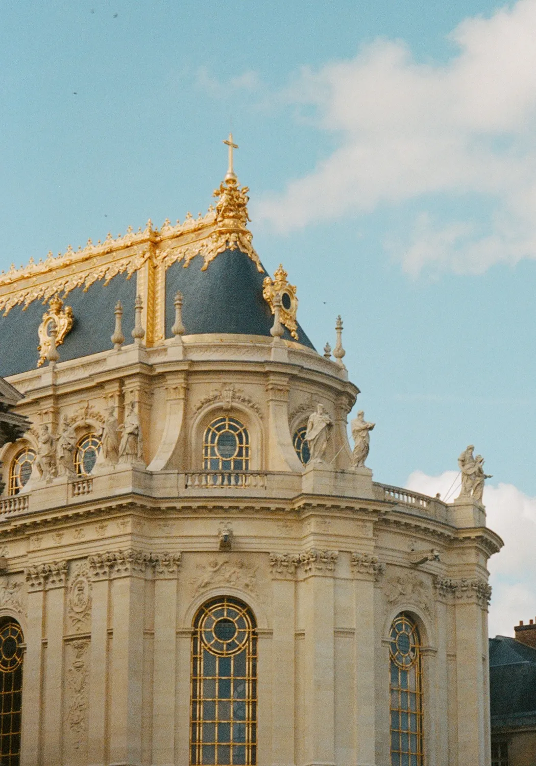 Ornate historic building corner topped with a dark slate roof decorated with gold trim and statues against a blue sky.
