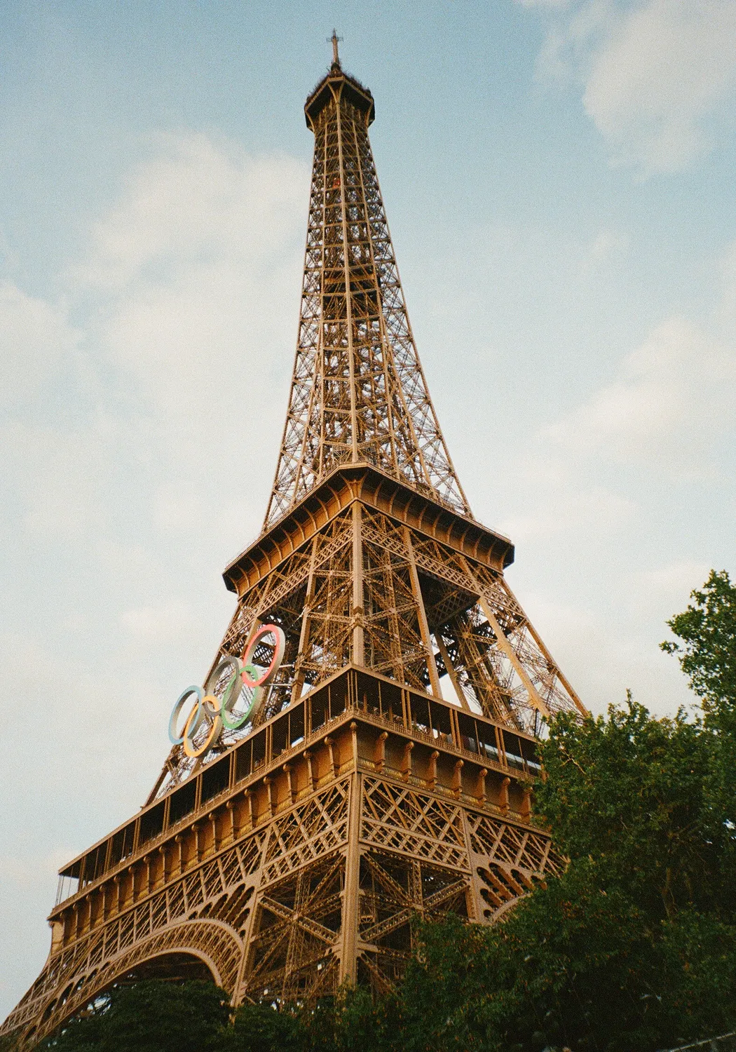 The Eiffel Tower in Paris with Olympic rings decoration and partly visible green trees under a cloudy sky.