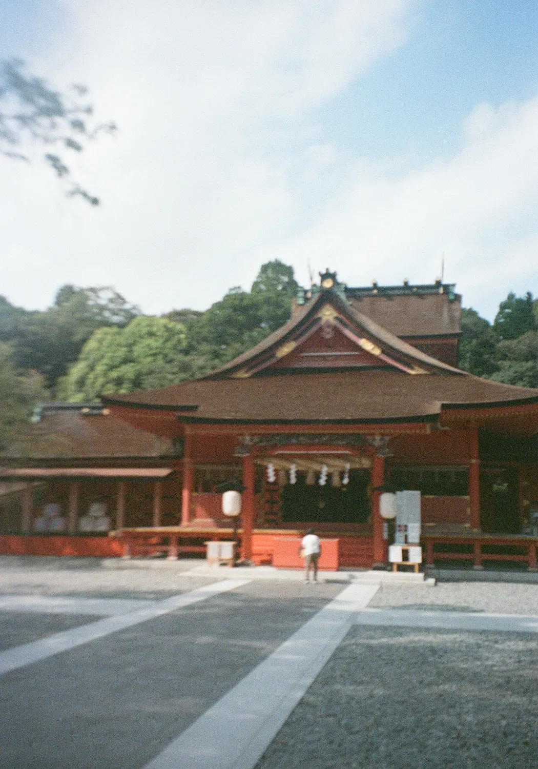 Traditional Japanese shrine with a person standing in front, surrounded by trees and a partly cloudy sky.