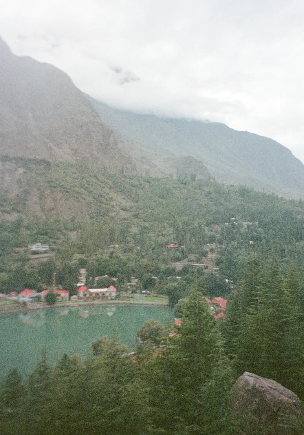 A serene mountain lake surrounded by dense green pine trees and small buildings with red roofs under a cloudy sky.