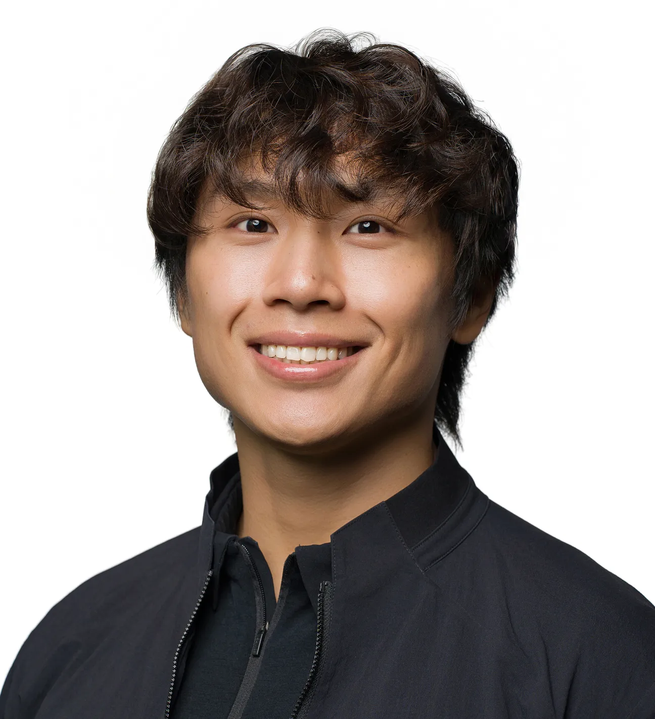 Smiling young man with wavy dark hair wearing a black jacket against a white background.