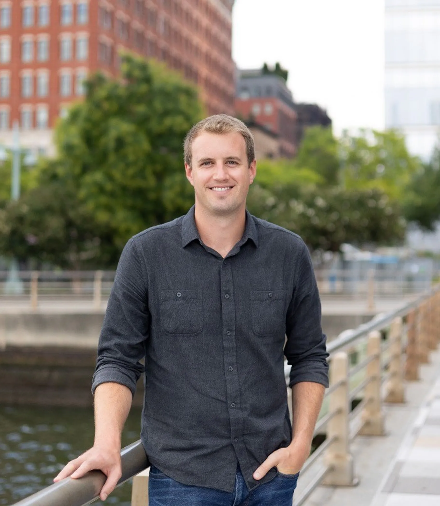 Smiling man in a dark gray shirt standing by a railing near water with urban buildings and trees in the background.