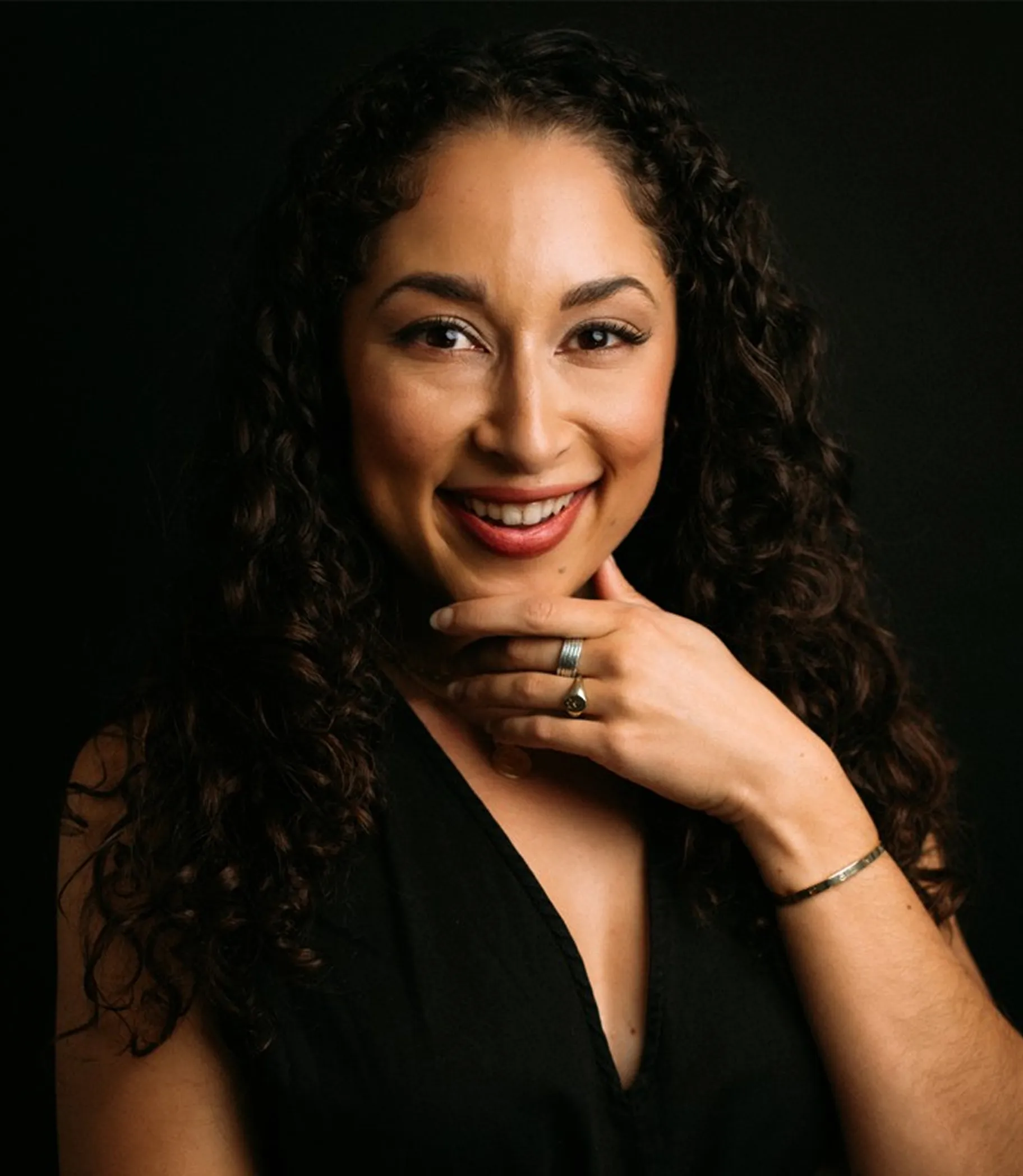 Smiling woman with curly dark hair wearing a black sleeveless top, resting her chin on her hand with rings and a bracelet.