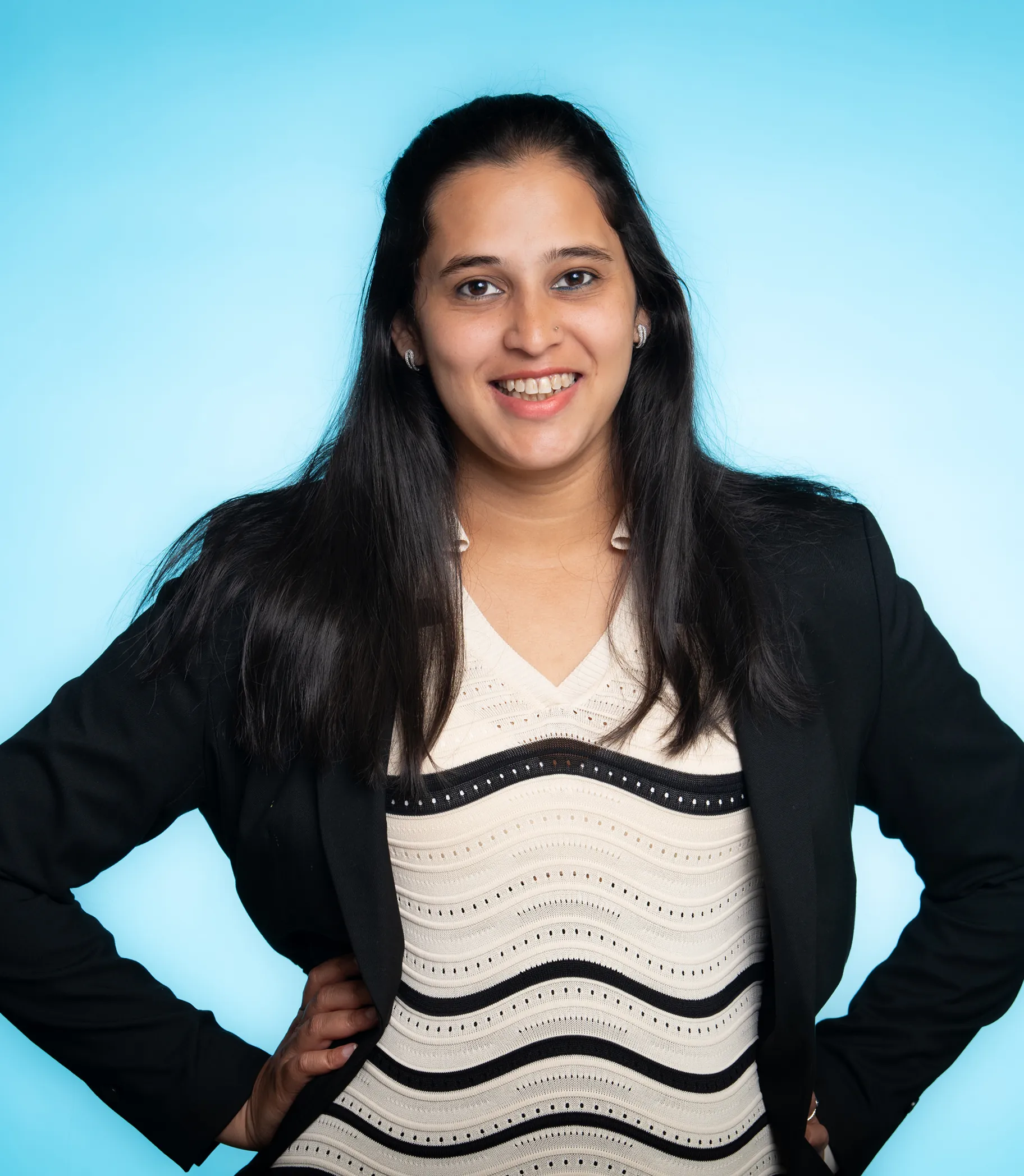 Smiling woman with long dark hair wearing a black blazer and a cream top with black wavy stripes against a light blue background.
